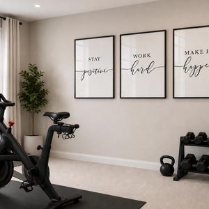 May include: A home gym featuring a black exercise bike, framed motivational art, a dumbbell rack, and a kettlebell. The art pieces read "Stay Positive", "Work Hard", and "Make It Happen" in elegant script.