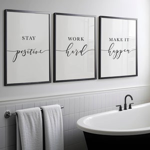 May include: Three framed wall art prints with motivational quotes: "Stay Positive," "Work Hard," and "Make It Happen." The prints have a minimalist design with black frames and white backgrounds. The bathroom setting includes a black bathtub and a towel rack.