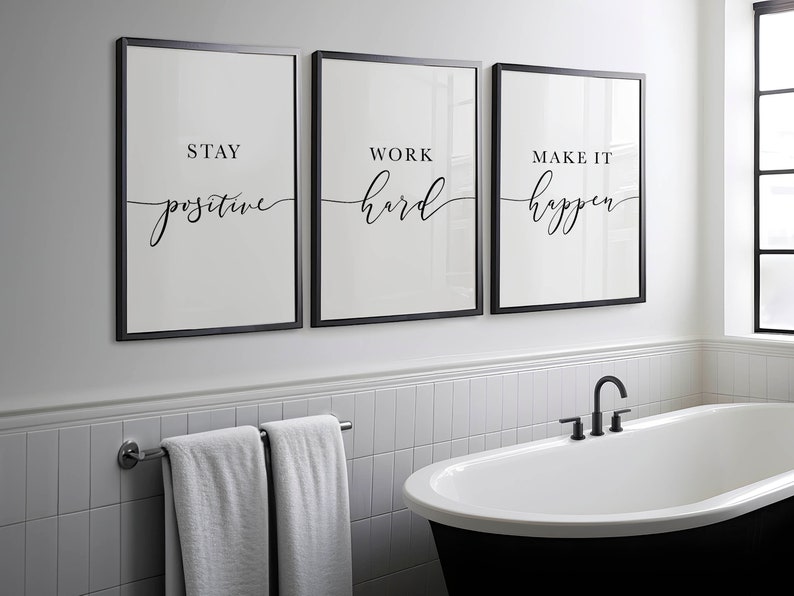 May include: Three framed wall art prints with motivational quotes: "Stay Positive," "Work Hard," and "Make It Happen." The prints have a minimalist design with black frames and white backgrounds. The bathroom setting includes a black bathtub and a towel rack.