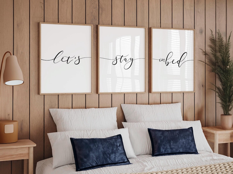 May include: Three framed prints with black calligraphy on a white background. The prints read "Let's stay in bed". The prints are hanging on a wood wall above a white bed with blue pillows.