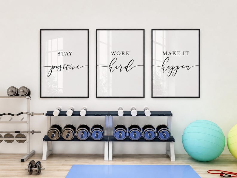 May include: Three framed motivational posters with black frames and white backgrounds. The posters read "Stay Positive", "Work Hard", and "Make it Happen" in elegant script. Dumbbells and exercise equipment are visible in the foreground.