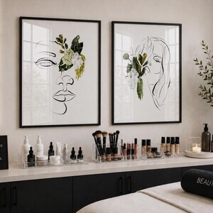 May include: Two framed art prints with black frames, featuring line drawings of faces adorned with floral elements. A black sign reads "CONFIDENCE IS BEAUTIFUL." Various beauty products are displayed on a white countertop.