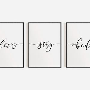 May include: Three black framed prints with white backgrounds and black calligraphy. The prints read "Let's", "stay", and "in bed".