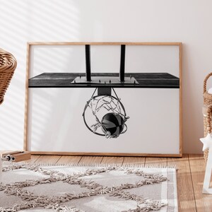 May include: Black and white framed print of a basketball going through a hoop. The image is a close-up shot of the basketball net and ball, with a wooden frame. The print is displayed in a room with a rug and wicker baskets.