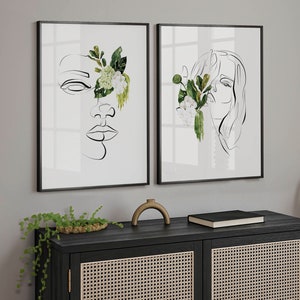 May include: Two framed art prints featuring minimalist line drawings of faces, each partially obscured by watercolor floral arrangements. The artwork is in black frames, and the floral elements include green leaves and white flowers. The prints are displayed on a gray wall.