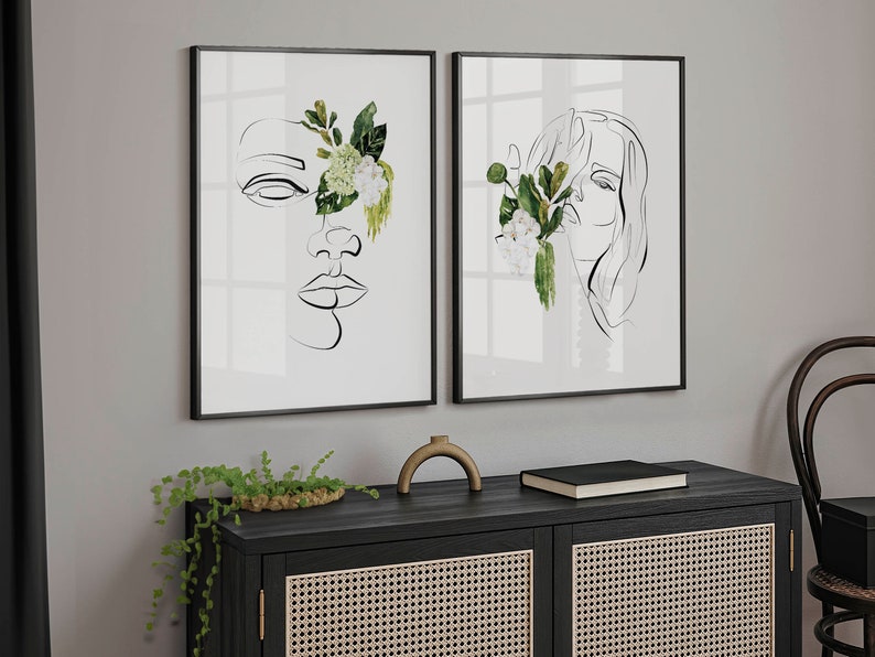 May include: Two framed art prints featuring minimalist line drawings of faces, each partially obscured by watercolor floral arrangements. The artwork is in black frames, and the floral elements include green leaves and white flowers. The prints are displayed on a gray wall.