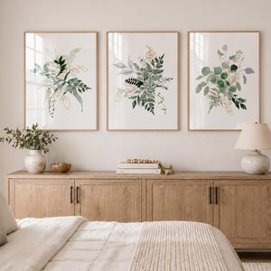 Puede incluir: Tres impresiones bot&aacute;nicas enmarcadas con arreglos de hojas verdes y doradas al estilo acuarela. Las impresiones se exhiben sobre un mueble de madera clara con una l&aacute;mpara, un jarr&oacute;n y objetos decorativos. La habitaci&oacute;n tiene una paleta de colores neutros.