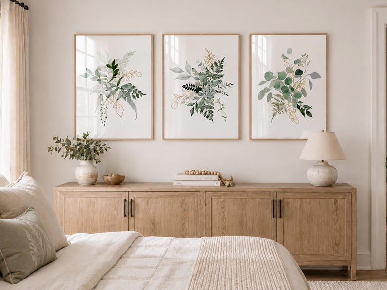 Puede incluir: Tres impresiones bot&aacute;nicas enmarcadas con arreglos de hojas verdes y doradas al estilo acuarela. Las impresiones se exhiben sobre un mueble de madera clara con una l&aacute;mpara, un jarr&oacute;n y objetos decorativos. La habitaci&oacute;n tiene una paleta de colores neutros.
