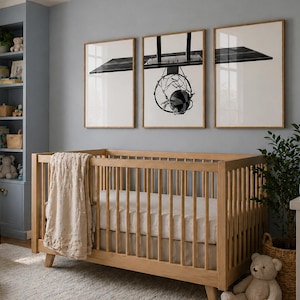 May include: A light wood crib with vertical slats and a beige textured blanket draped over the side. Above the crib, three framed prints display a black and white basketball hoop and backboard, with the hoop centered in the middle frame.