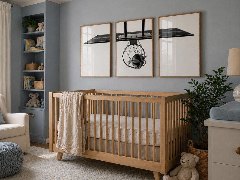 May include: A light wood crib with vertical slats and a beige textured blanket draped over the side. Above the crib, three framed prints display a black and white basketball hoop and backboard, with the hoop centered in the middle frame.