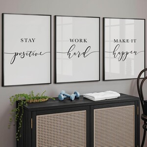 May include: Three framed motivational art prints with black frames and white backgrounds. The prints read "Stay Positive", "Work Hard", and "Make it Happen" in black script. A black cabinet with a woven front, dumbbells, and a towel are below.