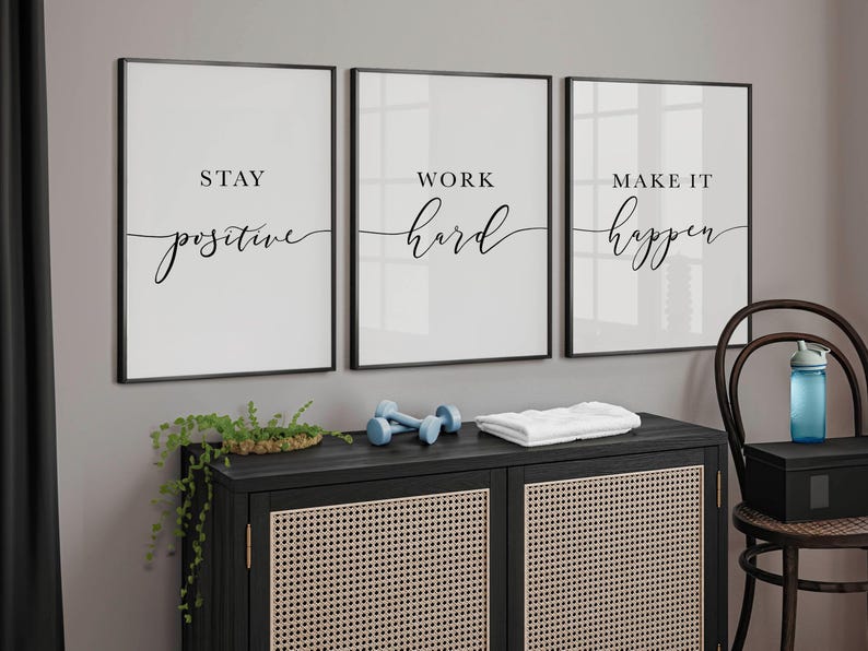 May include: Three framed motivational art prints with black frames and white backgrounds. The prints read "Stay Positive", "Work Hard", and "Make it Happen" in black script. A black cabinet with a woven front, dumbbells, and a towel are below.
