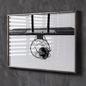 May include: Black and white framed photograph of a basketball hoop with a ball going through the net. The image is a close-up shot, with the hoop and ball in sharp focus. The background is blurred, with a window and blinds visible.