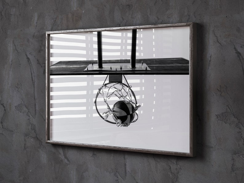 May include: Black and white framed photograph of a basketball hoop with a ball going through the net. The image is a close-up shot, with the hoop and ball in sharp focus. The background is blurred, with a window and blinds visible.
