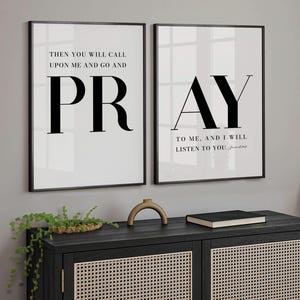 May include: Two framed art prints with black text on a white background. One reads "PRAY" and the other reads "Then you will call upon me and go and PR". The prints are displayed above a black cabinet.