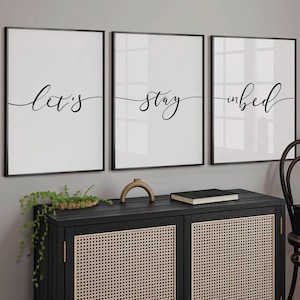 May include: Three black framed prints with white backgrounds and black calligraphy. The prints read "Let's stay in bed".