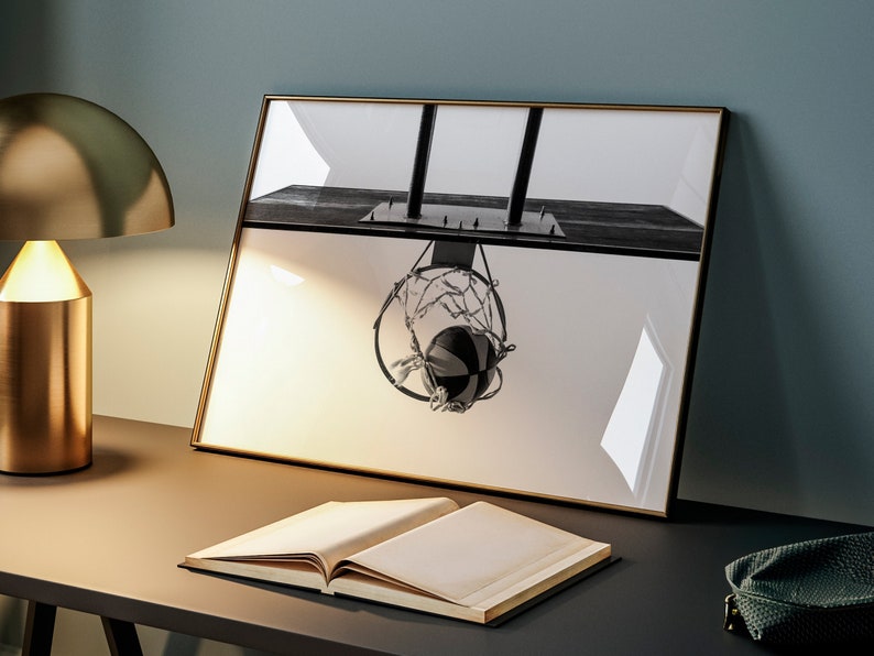May include: Black and white framed print of a basketball hoop with a ball in the net. The image is displayed on a desk with a gold lamp and an open book. The print has a gold frame.