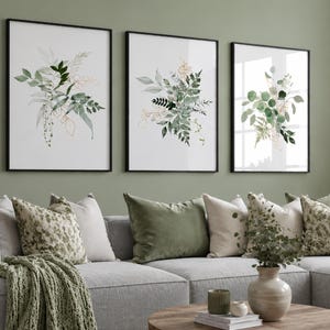 Puede incluir: Tres impresiones art&iacute;sticas bot&aacute;nicas enmarcadas con ilustraciones de acuarela de hojas verdes y doradas. Las impresiones est&aacute;n encima de un sof&aacute; gris con cojines decorativos y una manta verde. Una mesa de centro redonda de madera est&aacute; frente al sof&aacute;.