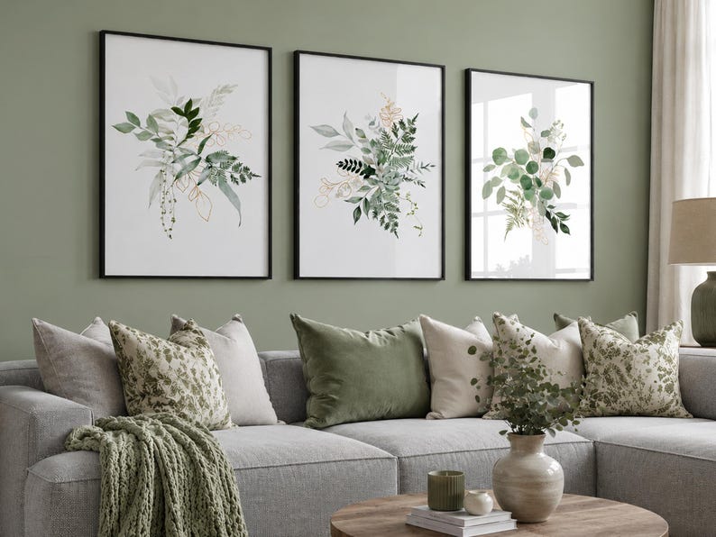 Puede incluir: Tres impresiones art&iacute;sticas bot&aacute;nicas enmarcadas con ilustraciones de acuarela de hojas verdes y doradas. Las impresiones est&aacute;n encima de un sof&aacute; gris con cojines decorativos y una manta verde. Una mesa de centro redonda de madera est&aacute; frente al sof&aacute;.