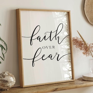 May include: A framed print with the words "faith over fear" in elegant black script. The artwork is displayed on a wooden shelf, with decorative items like a vase and a small, round, textured object. The frame is a light wood color.