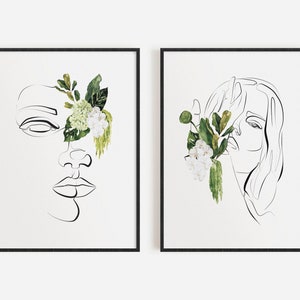 May include: Two framed art prints featuring minimalist line drawings of faces. One face has a closed eye and is partially adorned with green leaves and white flowers. The other print shows a face with long hair, also with floral accents. Both are in black frames.