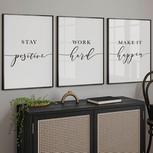 May include: Three framed motivational art prints with black frames and white backgrounds. The prints read "Stay Positive", "Work Hard", and "Make It Happen" in black script. The prints are displayed above a black cabinet with woven doors.