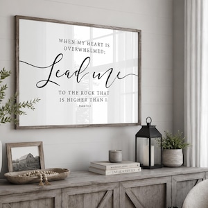 Op de afbeelding: Een rechthoekig wandbord met een donkerhouten lijst, een witte achtergrond en zwarte tekst: WHEN MY HEART IS OVERWHELMED; Lead me TO THE ROCK THAT IS HIGHER THAN I. Psalm 61:2. De typografie combineert een schreeflettertype met een scriptlettertype.
