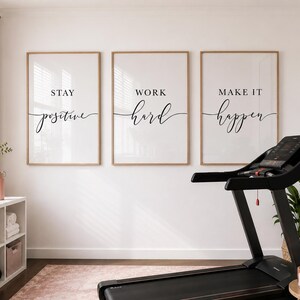 May include: A home gym scene featuring three framed prints with motivational phrases: "Stay Positive," "Work Hard," and "Make It Happen." A black treadmill is in the foreground, with a pink rug and decor adding a touch of color. A mirror and plant complete the space.