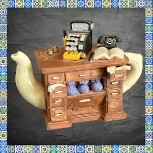 May include: A novelty teapot shaped like a wooden desk with drawers, featuring a black cash register, a black rotary telephone, an open book, and gold coins on top. Three blue money bags are visible in the centre open shelf of the desk.