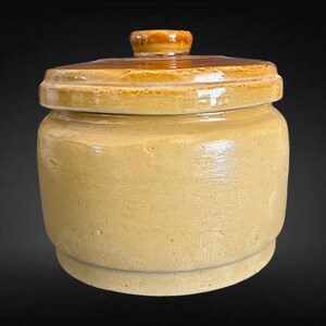 May include: A tan ceramic jar with a lid. The jar has a rounded body and a lid with a small knob. The lid and knob are a darker brown colour. The jar is likely vintage or antique.