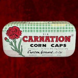 May include: A vintage "Carnation Corn Caps" tin featuring a red carnation illustration. The tin has a green and white checkered pattern on the top and red stripes at the bottom. The text includes "Cuxson, Gerrard & Co. Ltd."