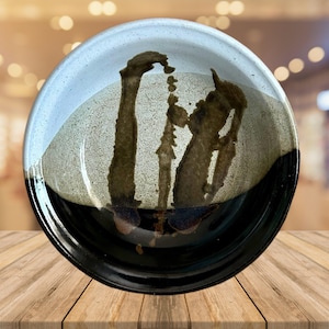 May include: A handcrafted ceramic bowl with a distinctive design. It has a black rim, a light blue upper section, and a speckled tan centre with dark brown abstract patterns. The bowl is displayed on a wooden surface, highlighting its artistic appeal.