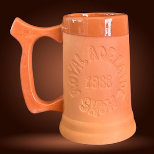 May include: A terracotta beer stein with a large handle. The stein features the embossed text "Royal Adelaide Show 1988". The handle and rim are a slightly darker shade of orange. A vintage drinking vessel.