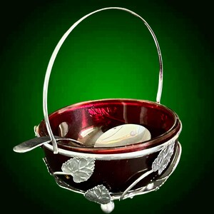 May include: A small, ruby red glass serving bowl with a silver handle and leaf detailing. The bowl contains a spoon and sits on a silver base. The handle arches over the bowl. The background is a dark green.