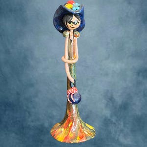 May include: A ceramic figurine of a woman wearing a blue hat decorated with colourful flowers. The figurine has a long, slender body with a small purse and a flared base, painted in vibrant hues of red, yellow, and orange.