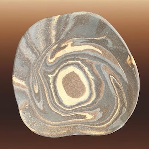 May include: A hand-formed ceramic plate with an irregular, organic shape. The surface features a swirling, marbled pattern in shades of charcoal grey, cream, and earthy tan, creating a continuous spiral effect that radiates from the centre.