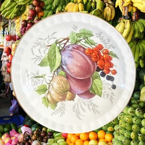 May include: A white ceramic plate with a painted design of fruit and leaves. The plate features plums, grapes, and other fruits in shades of purple, red, and green.