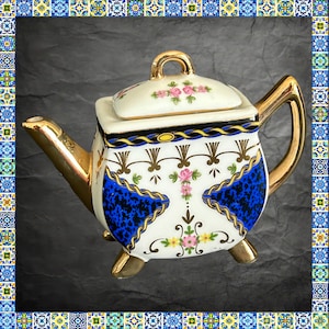 May include: A square-shaped ceramic teapot with a lid, featuring a white base with blue and black mottled panels, gold trim on the handle, spout, and feet, and floral motifs in pink and yellow with gold filigree accents throughout the body.