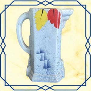 May include: A light blue ceramic pitcher with a speckled finish, featuring a curved handle and a spout. The front is decorated with a yellow leaf, a red leaf, and a blue geometric staircase pattern. The pitcher has a square base and body.