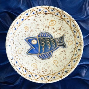 May include: A ceramic plate with a hand-painted design featuring a blue fish in the centre. The plate has a speckled off-white background with a decorative border of brown and blue dots and swirls. The fish has detailed scales and an open mouth.