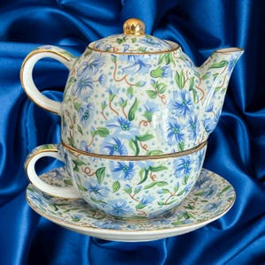 May include: A floral patterned tea set comprising a teapot, teacup, and saucer. The design features blue flowers, green leaves, and gold detailing. The teapot has a gold knob. The teacup is placed on the saucer.