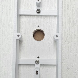 May include: A white rectangular electrical box with a central circular opening and screw holes. The box has a grid-like structure and is mounted on a textured white surface. The design suggests it is for electrical components.