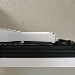 May include: A white wall-mounted bracket with a black and white device attached. The bracket is mounted on a textured wall. The device has a tiered, black, circular design.