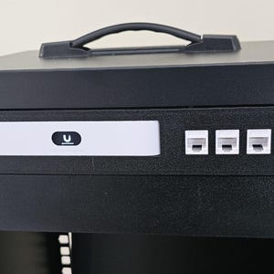 Peut inclure: Rack réseau noir avec une poignée sur le dessus. Le panneau avant comporte un appareil blanc avec un logo 'U' et trois ports RJ45 blancs. Le rack est fixé avec des vis noires.