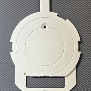 Könnte beinhalten: Ein weißes Kunststoffteil, kreisförmig mit einem rechteckigen Sockel und einer kleinen Lasche oben. Der kreisförmige Teil hat konzentrische Ringe und ein zentrales Loch. Der Sockel hat ebenfalls ein Loch. Auf grauem, strukturiertem Hintergrund.