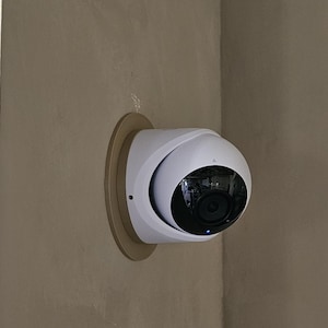 May include: A white security camera with a black lens is mounted on a beige wall. The camera is attached to a circular, light brown mounting plate. The camera has a rounded design and a small blue light.