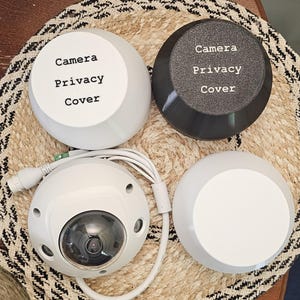 May include: A white security camera with a clear dome lens, accompanied by two white and one dark gray camera privacy covers. The covers have the text "Camera Privacy Cover" printed on them. The camera has a white cable.