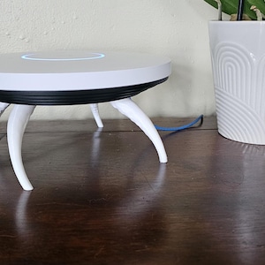 May include: A white and black electronic device with curved white legs, resembling a small table. A blue cable extends from the device. A white, patterned flower pot with a plant is to the right.