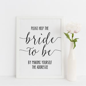 May include: A framed white sign with black script that reads "PLEASE HELP THE bride TO BE BY MAKING YOURSELF THE ADDRESSEE." A white vase holds a white peony flower. The background is white.