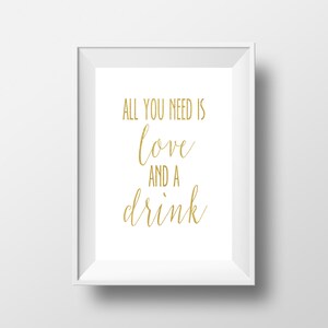 May include: A framed print with the words "ALL YOU NEED IS love AND A drink" in gold script on a white background. The print is in a white frame and is displayed against a white wall.